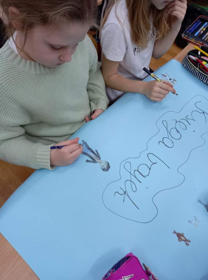 Zdjęcie przedstawia dwie uczennice w trakcie rysowania na plakacie – dużej karcie papieru leżącej na ławce szkolnej