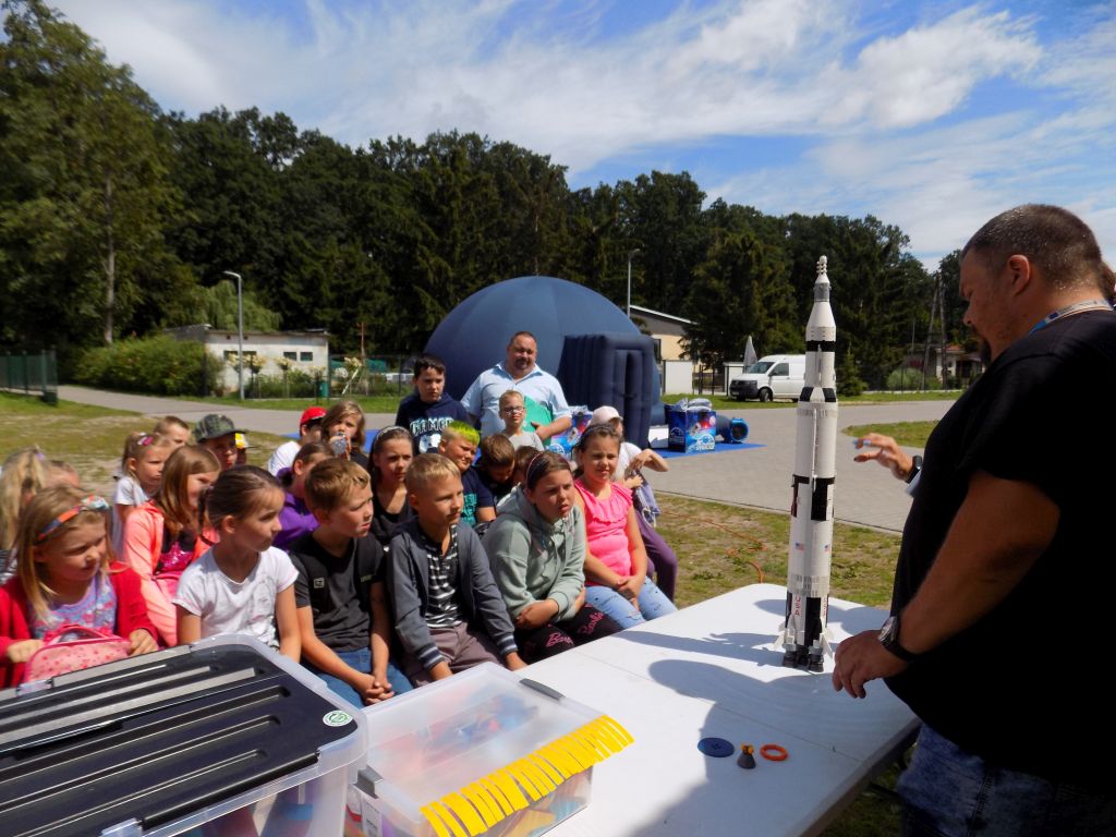 Fotografia przedstawia uczestników zajęć obserwujących budowę rakiety. W tle widać namiot, a rakieta jest bardzo wysoka