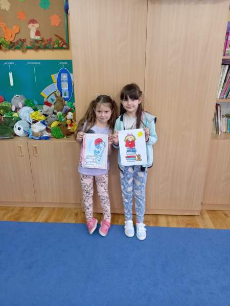 Na zdjęciu są dwie dziewczynki które prezentują swoje kolorowe rysunki. Na ścianie wiszą ozdoby jesienne