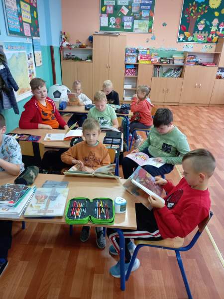 Na zdjęciu widać grupkę dzieci siedzących przy stolikach i czytających różne bajeczki o tematyce jesiennej. W tle na ścianie wiszą dekoracje jesienne