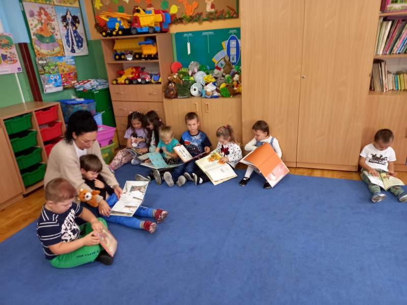 Na zdjęciu mamy grupę dzieci wraz z ich opiekunką siedzących wspólnie na wykładzinie. Czytają razem książki.