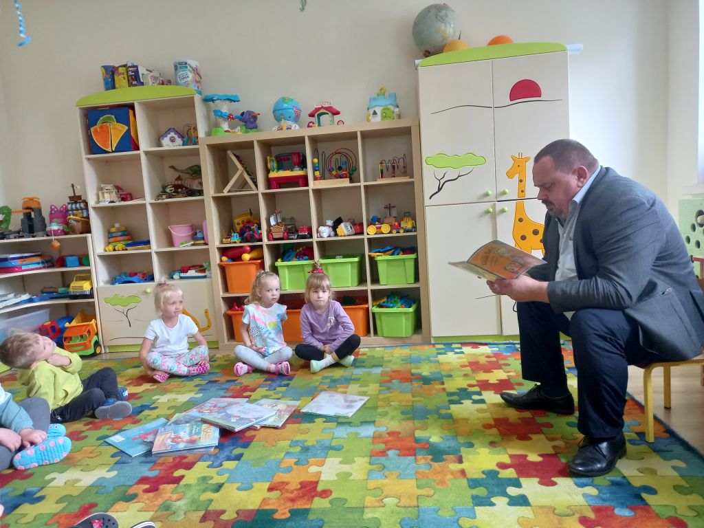 Fotografia przedstawia dyrektora biblioteki czytającego dzieciom bajkę. Dzieci siedzą w przedszkolu na dywanie przed dyrektorem. Dyrektor siedzi na krześle