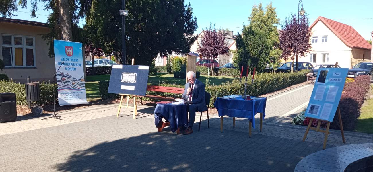 Zdjęcie przedstawia Burmistrza Miasta i Gminy Skępe Piotra Wojciechowskiego siedzącego przy stoliku i czytającego jeden z wierszy Adama Mickiewicza