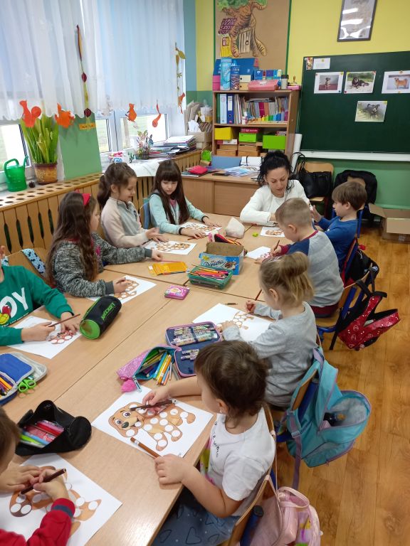 Fotografia przedstawia dzieci siedzące w szkolnych ławkach w trakcie lekcji kolorujące szablony na kartkach papieru