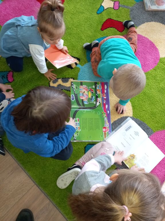 Zdjęcie przedstawia dzieci siedzące na dywanie w przedszkolu. Dzieci przeglądają kolorowe książeczki
