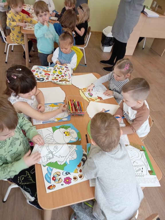 Zdjęcie przedstawia dzieci w przedszkolu siedzące przy stolikach w trakcie zajęć . Dzieci uzupełniają ilustracje w książkach naklejkami