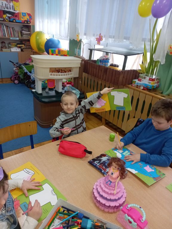 Zdjęcie przedstawia dzieci siedzące w klasie w ławkach w trakcie zajęć . Dzieci kolorują szablony książek dla dzieci