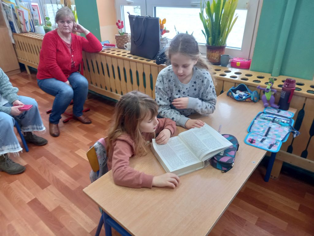 Zdjęcie przedstawia dwójkę dzieci siedzących w klasie w ławce szkolnej. Dzieci przeglądają gruby słownik