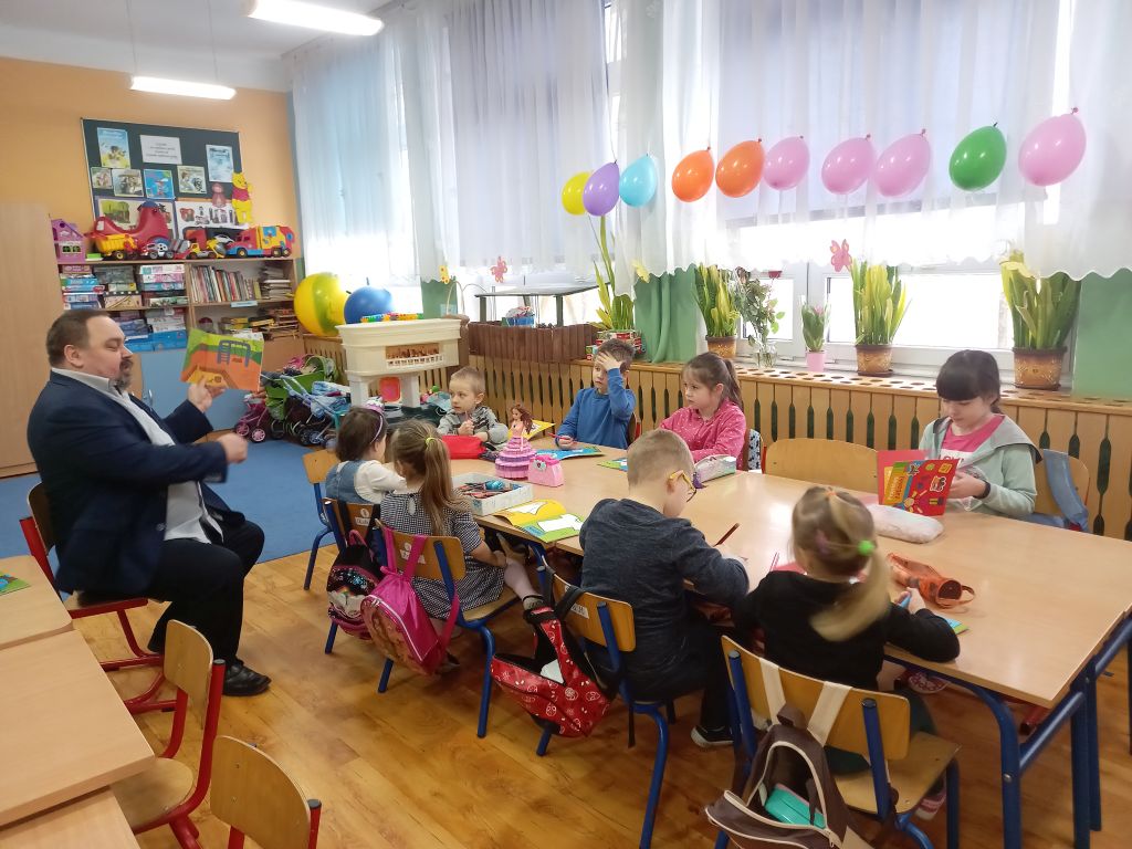 Zdjęcie przedstawia dyrektora biblioteki siedzącego w klasie przed dziećmi w trakcie prowadzenia zajęć lekcyjnych