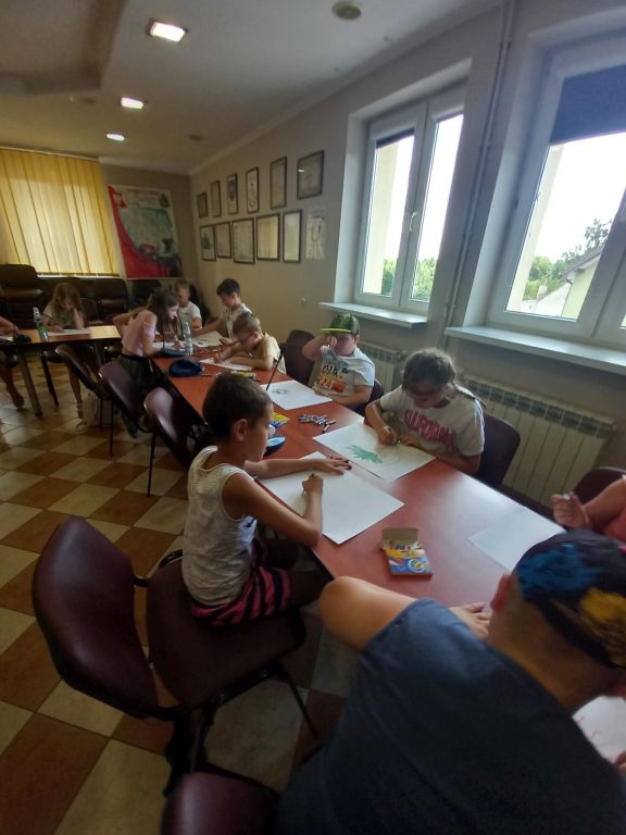 Zdjęcie przedstawia dzieci siedzące przy stolikach w Sali konferencyjnej urzędu w trakcie zajęć plastycznych