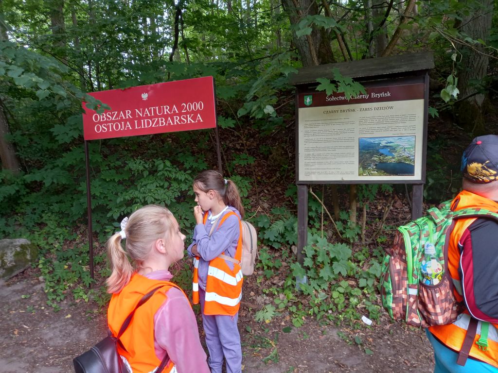 Zdjęcie przedstawia grupkę dzieci stojących w w parku krajobrazowym w Ośrodku „Wilga”. W tle widać napis Obszar Natura 2000 Ostoja Lidzbarska