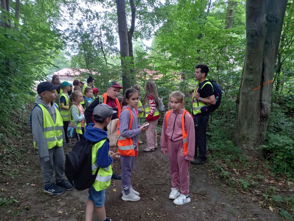 Zdjęcie przedstawia grupkę dzieci stojących w w parku krajobrazowym w Ośrodku „Wilga” w Górznie. Przed dziećmi stoi przewodnik opowiadający im o Ośrodku