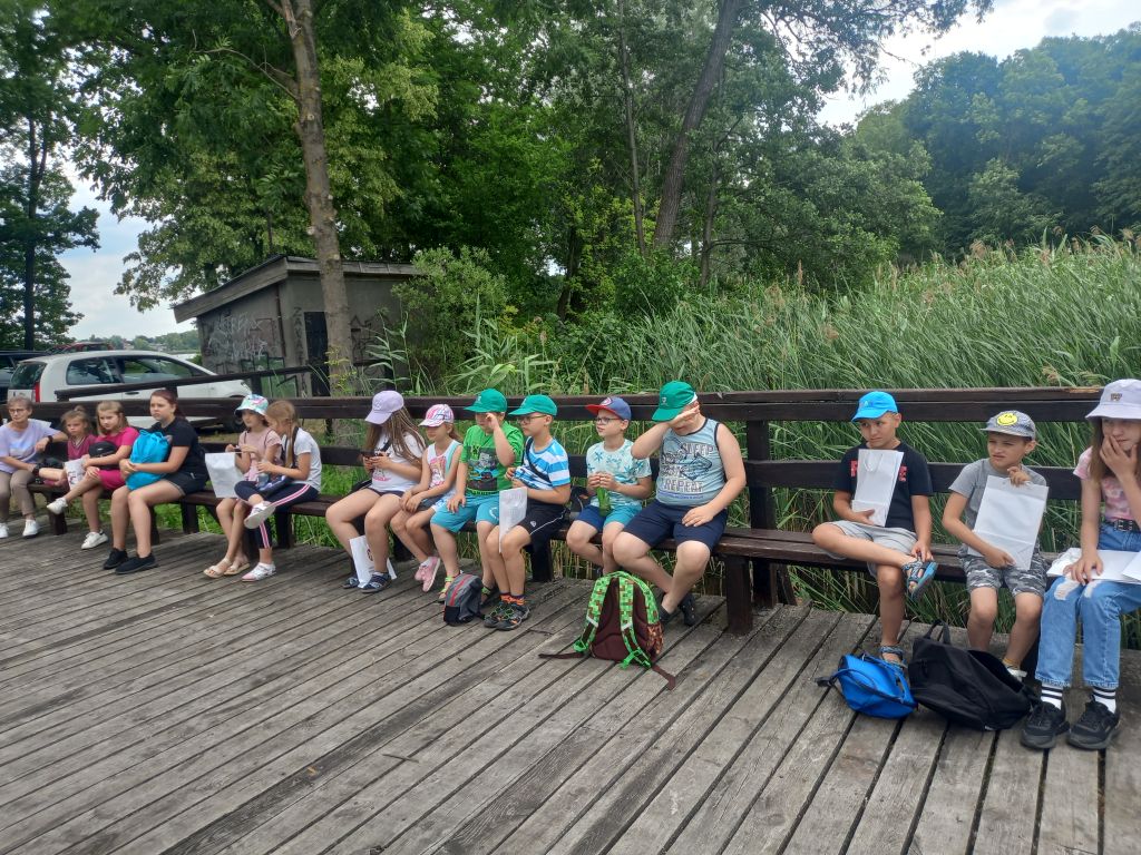 Zdjęcie przedstawia grupkę dzieci siedzących na ławce na pomoście nad jeziorem