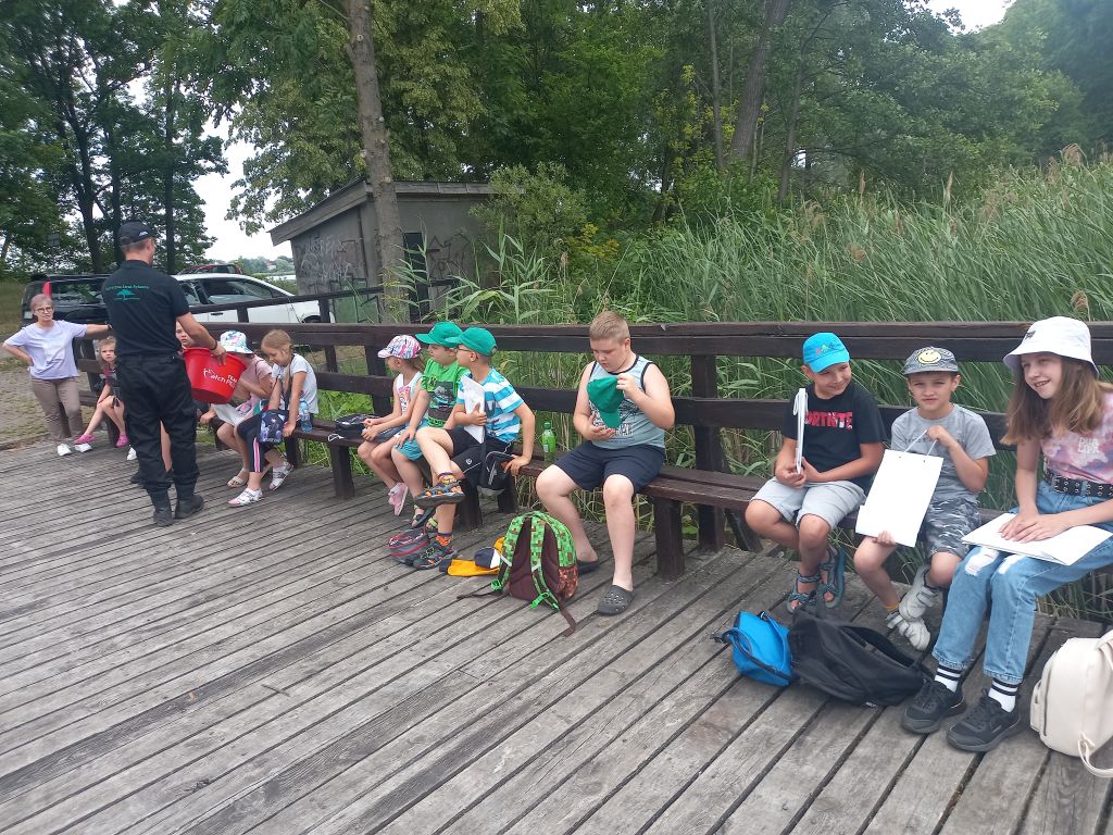 Zdjęcie przedstawia grupkę dzieci siedzących na ławce na pomoście nad jeziorem. Przed dziećmi stoi wędkarz pokazujący dzieciom akcesoria do wędkowania