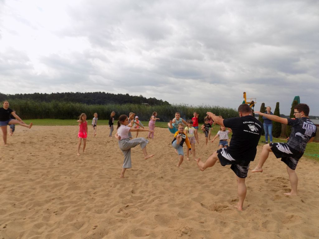 Zdjęcie przedstawia uczestników zajęć w trakcie zajęć w ramach sztuk walki na boisku do piłki plażowej nad jeziorem