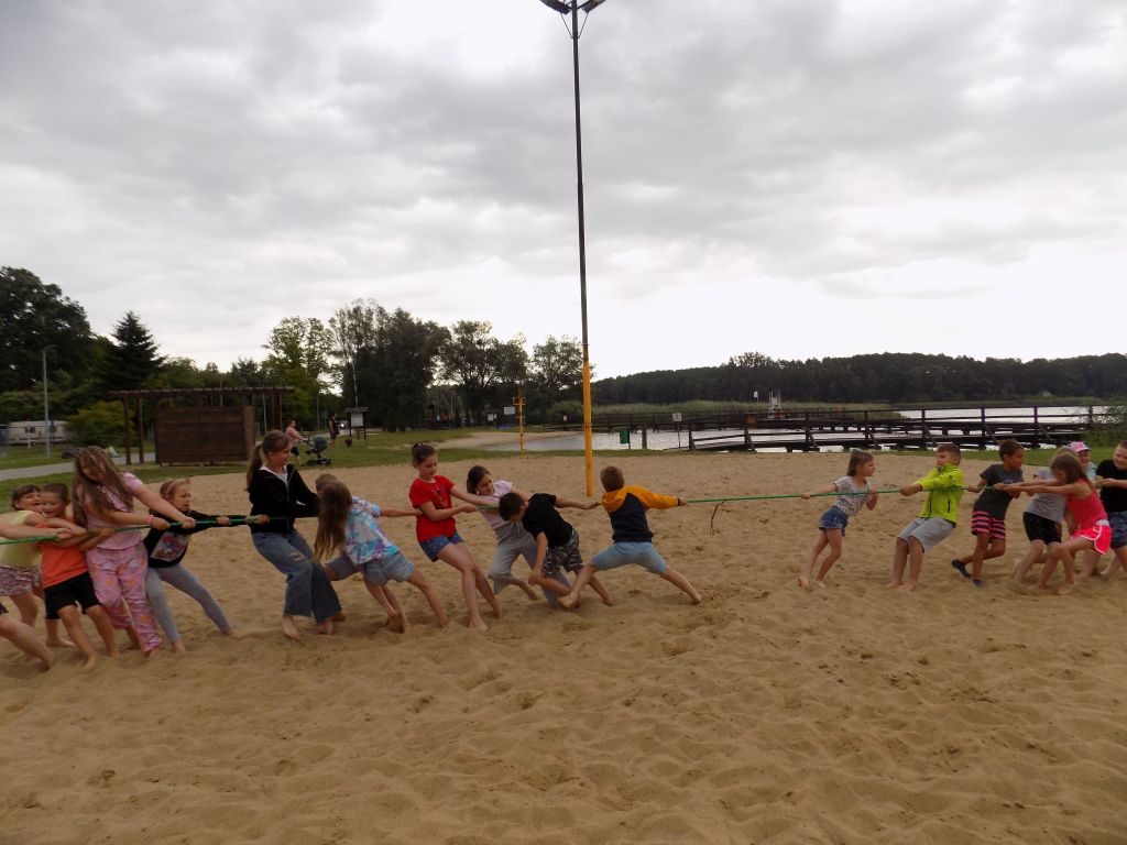 Zdjęcie przedstawia uczestników zajęć w trakcie zajęć w ramach sztuk walki na boisku do piłki plażowej nad jeziorem w trakcie przeciągania liny