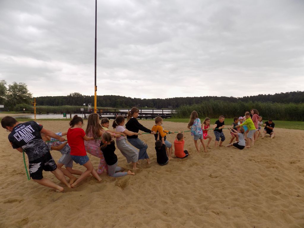 Zdjęcie przedstawia uczestników zajęć w trakcie zajęć w ramach sztuk walki na boisku do piłki plażowej nad jeziorem w trakcie przeciągania liny