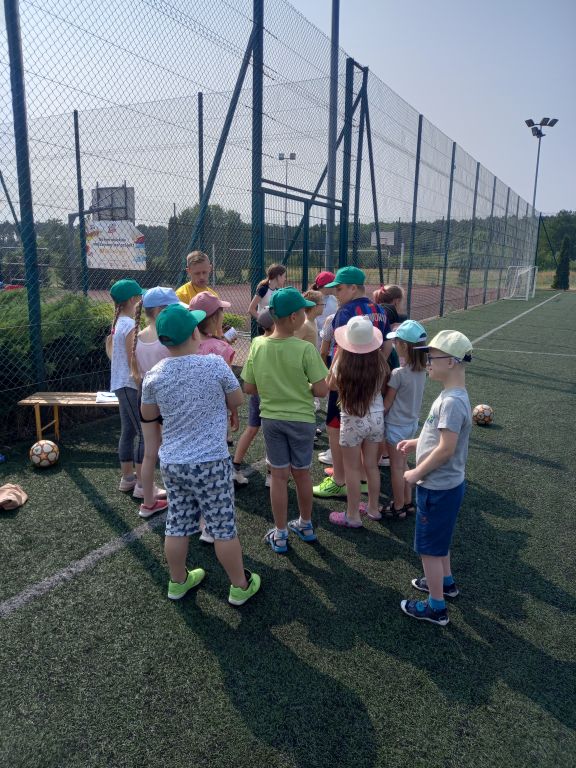 Zdjęcie przedstawia uczestników zajęć wakacyjnych w trakcie zbiórki na rozpoczęcie zabaw na Orliku. Dzieci stoją ustawione wokół trenera
