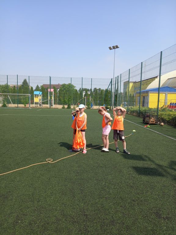 Zdjęcie przedstawia grupkę dzieci w pomarańczowych kamizelkach stojących na boisku orlik w trakcie zabawy