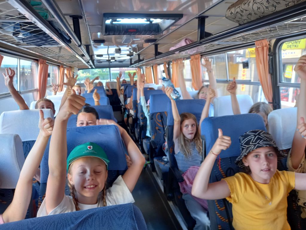 Zdjęcie przedstawia dzieci siedzące w autobusie z radośnie uniesionymi rękami