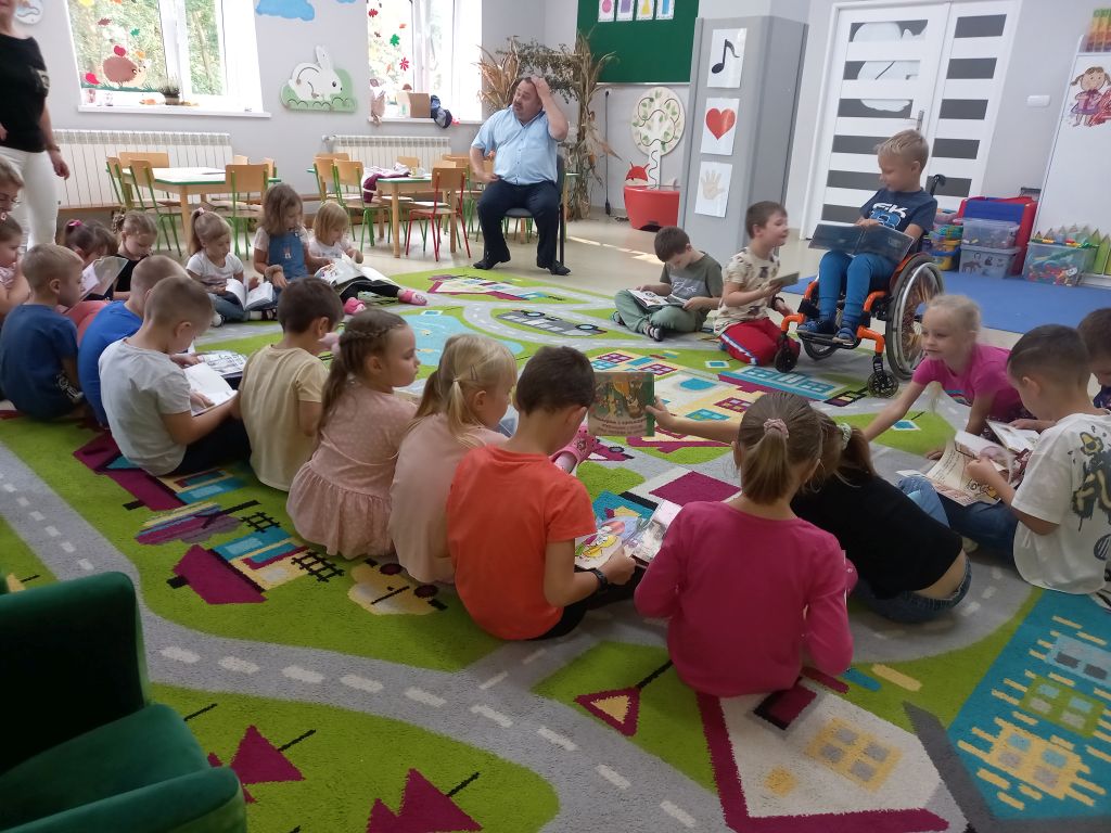 Na zdjęciu widać grupkę dzieci w siedzących na dywanie w przedszkolu. Przed dziećmi na krześle siedzi dyrektor biblioteki