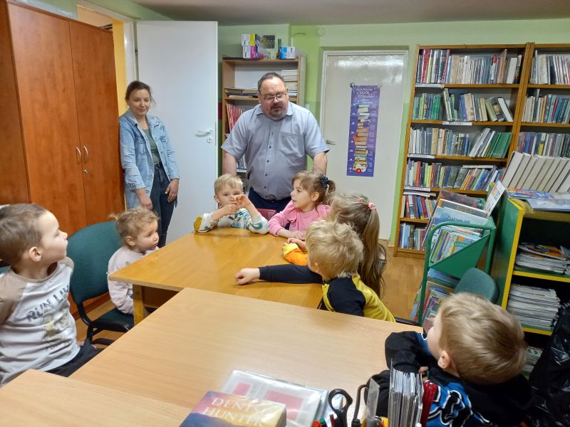 Na zdjęciu widać dzieci siedzące w bibliotece przy stolikach i dyrektora biblioteki stojącego przed dziećmi w trakcie prowadzonych zajęć