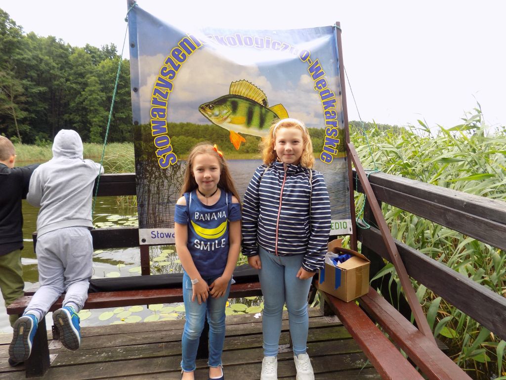 dziewczynki stojące na pomoście na tle baneru z logiem Stowarzyszenia Ekologiczno Wędkarskiego Okoń