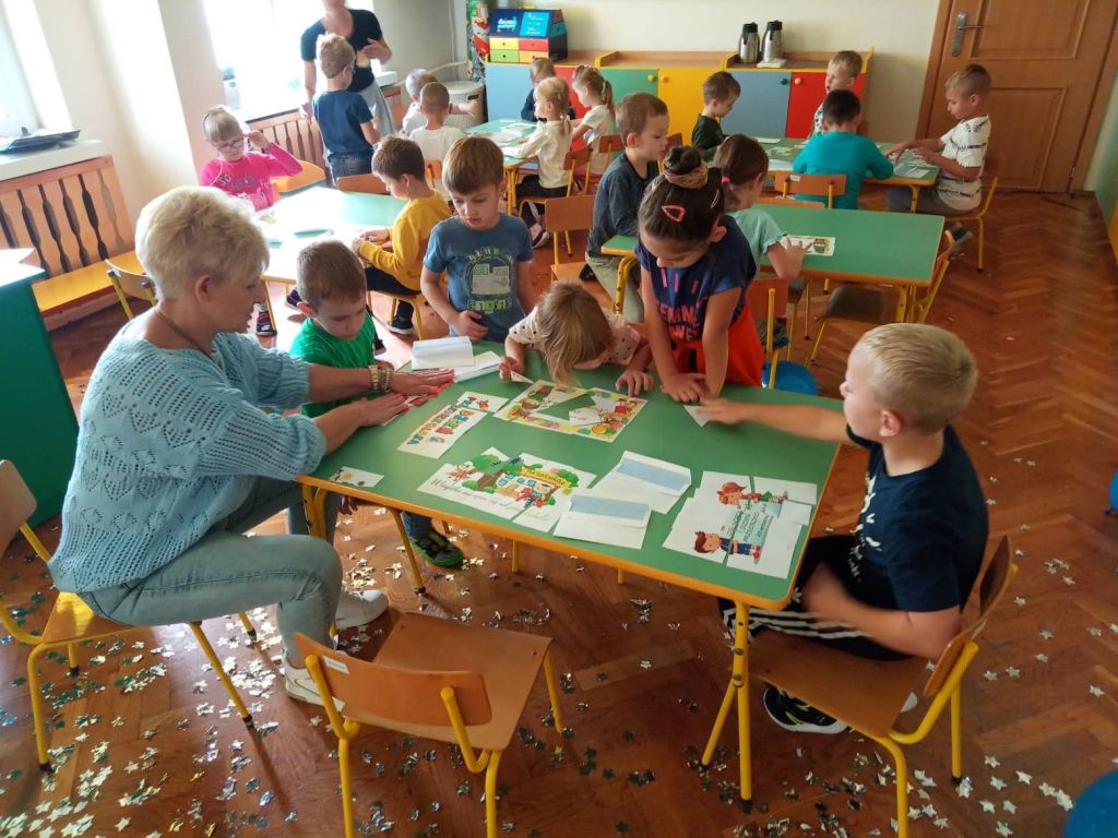 Na fotografii widać dzieci, które wraz z dyrektorem Przedszkola Panią Iwoną Małkiewicz układają puzzle na stolikach