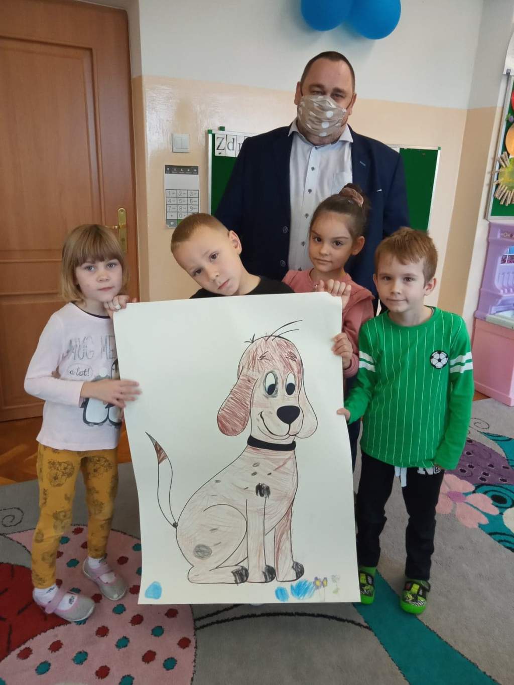 Zdjęcie przedstawia dyrektora biblioteki i czwórkę przedszkolaków w przedszkolu trzymających dużą kartkę z pokolorowanym rysunkiem psa