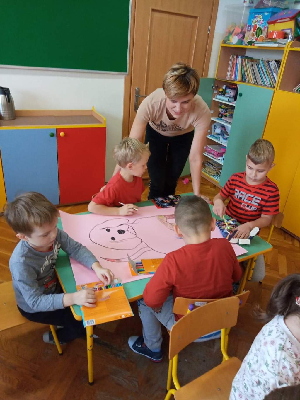 Zdjęcie przedstawia trójkę dzieci siedzących przy stoliku w przedszkolu i kolorujących obrazek pieska narysowany na dużej karcie papieru