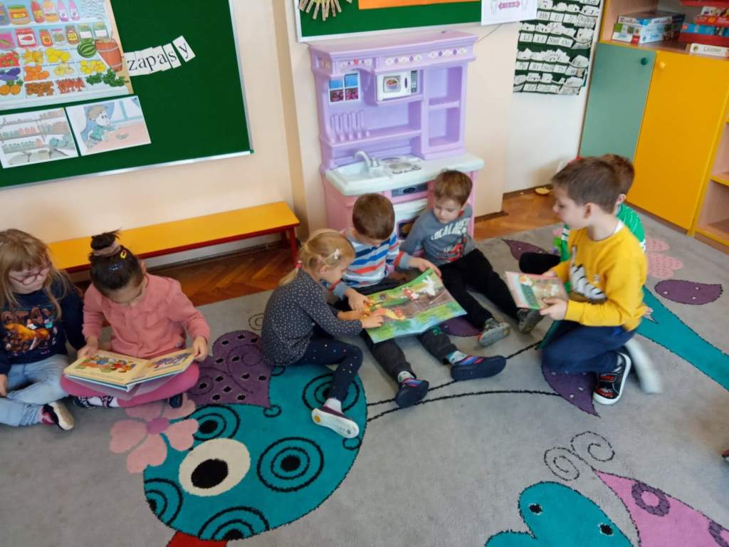 Zdjęcie przedstawia grupkę dzieci wraz z przedszkolankami. Wszyscy wycinają nożyczkami z papieru zakładki do ksiązek