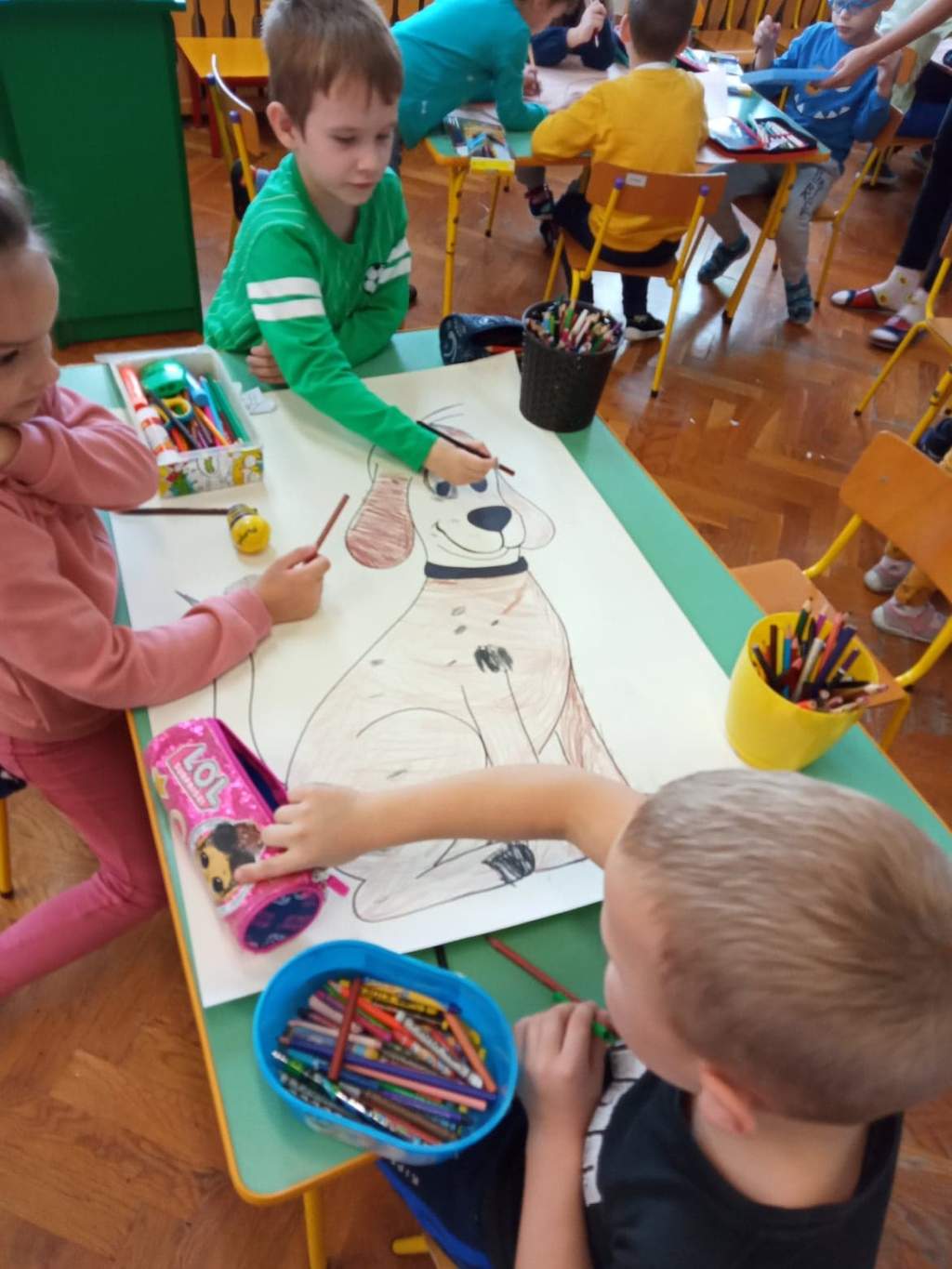 Zdjęcie przedstawia czwórkę dzieci siedzących przy stoliku w przedszkolu i kolorujących obrazek pieska narysowany na dużej karcie papieru