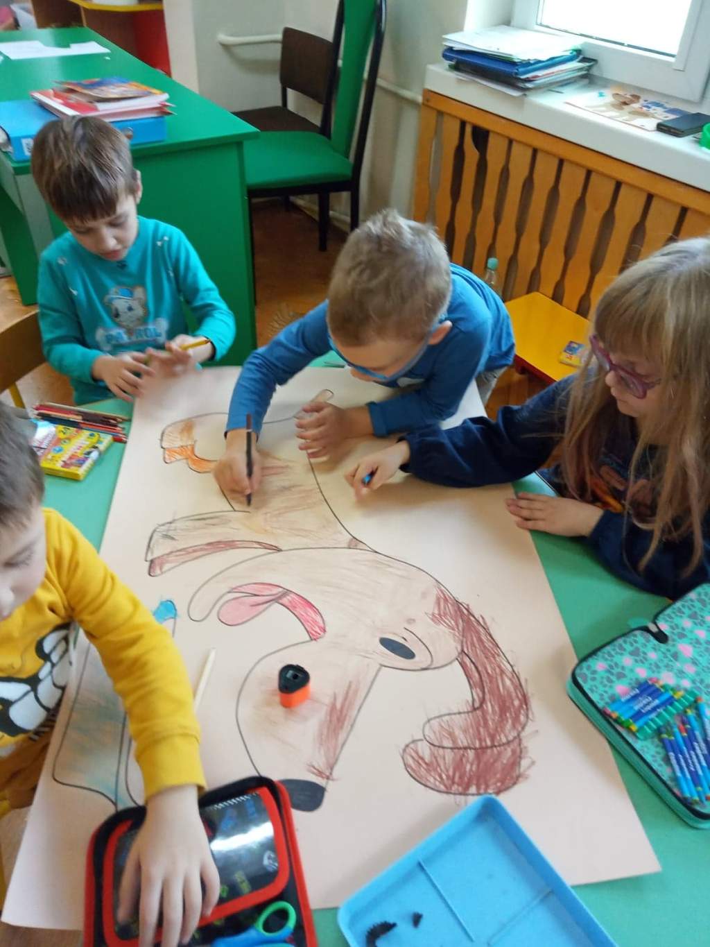 Zdjęcie przedstawia czwórkę dzieci siedzących przy stoliku w przedszkolu i kolorujących obrazek pieska narysowany na dużej karcie papieru. Nad dziećmi pochyla się nauczyczycielka i patrzy na obrazek.