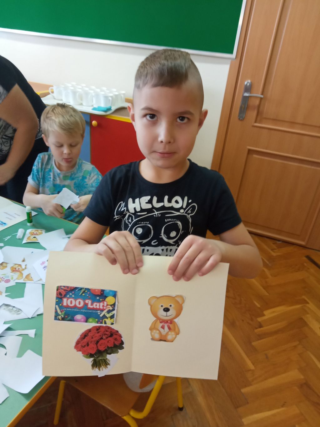 Zdjęcie przedstawia chłopca stojącego obok stolika w przedszkolu. Chłopiec trzyma w rękach wykonana przez siebie laurkę. W tle widać kolejnego chłopca wycinającego obrazki z kartki papieru.