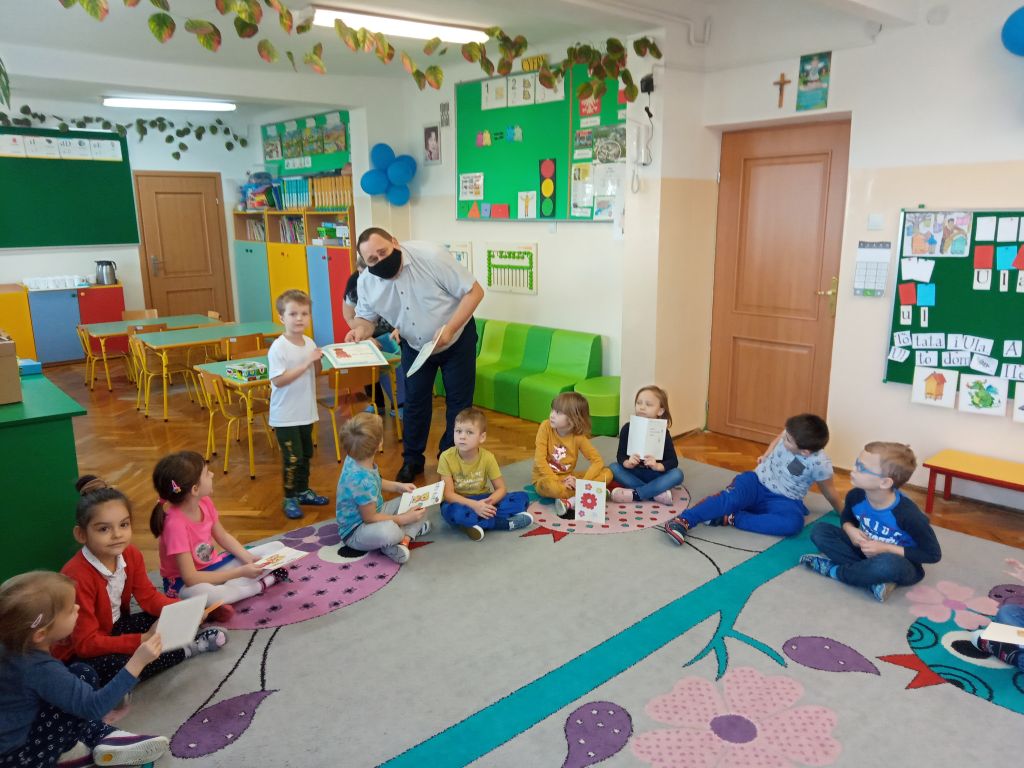 Na fotografii widać grupkę dzieci siedzących w kręgu na dywanie w przedszkolu. w tle widać dyrektora biblioteki wręczającego dyplom przedszkolakowi.