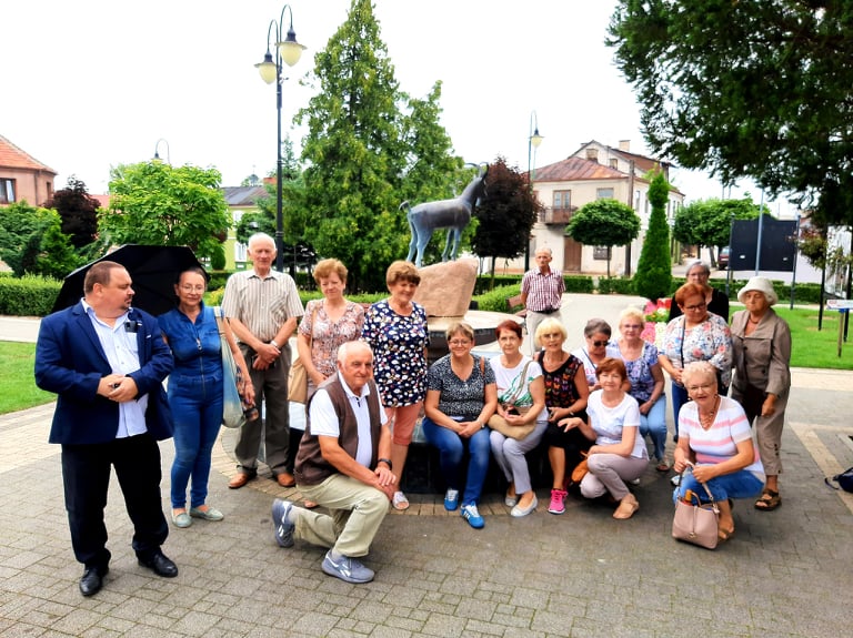 Na zdjęciu widać uczestników wycieczki i dyrektora biblioteki w Skępem pozujących do grupowego zdjęcia przy fontannie z koziołkiem w centrum miasta