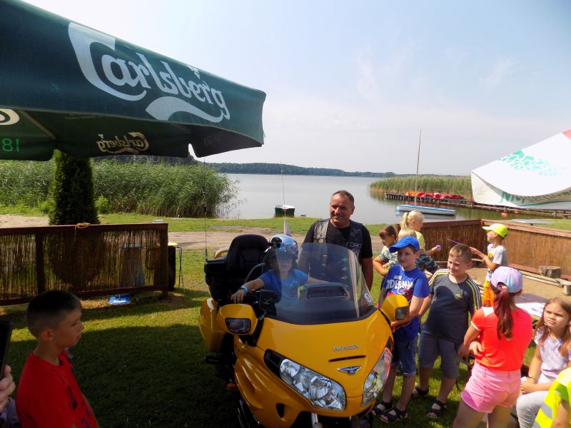 Na zdjęciu widać grupkę dzieci stojących nad jeziorem wraz z przedstawicielem  Klubu Motorowego „Wolny Duch” zgromadzonych wokół  motocykla