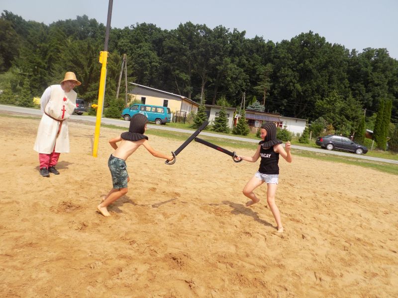 Na zdjęciu na pierwszym planie widać dwoje dzieci walczących na miecze wykonane z gąbki . Obok stoi pracownik muzeum przebrany w strój średniowieczny. Walka odbywa się na piasku na boisku do piłki siatkowej