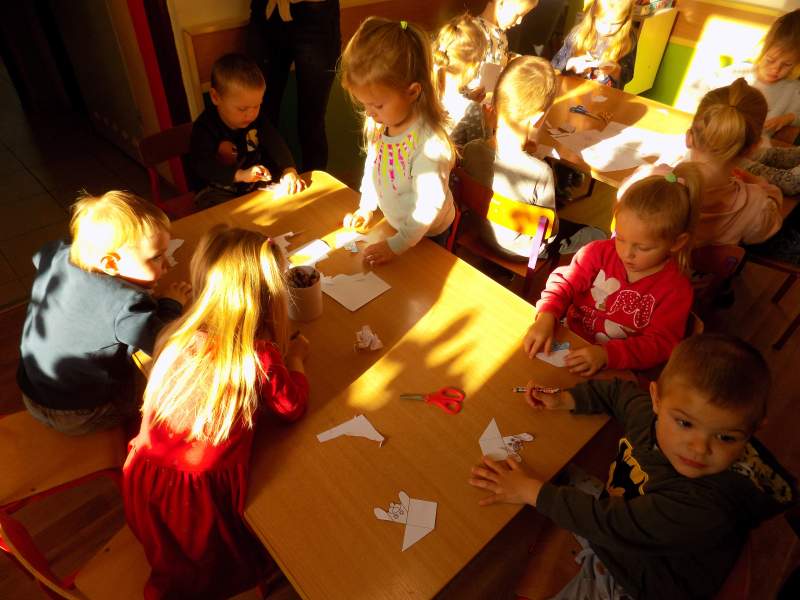 Na zdjęciu widać dzieci siedzące przy stoliku i wycinające z kartek papieru zakładki do książek