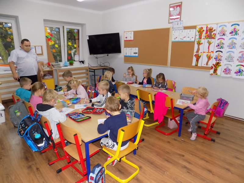 Na zdjęciu widać dzieci siedzące przy stolikach w trakcie kolorowania obrazków. W tle widać dyrektora Biblioteki