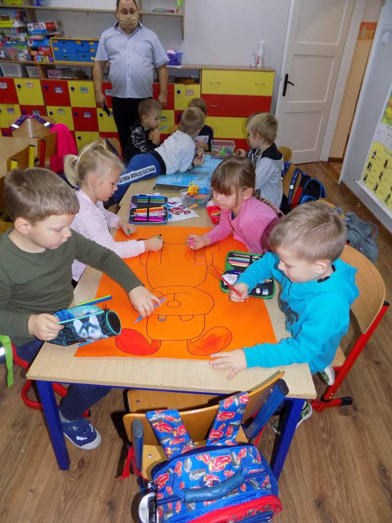 Na zdjęciu widać dzieci siedzące przy stolikach w trakcie kolorowania obrazków. W tle widać dyrektora Biblioteki