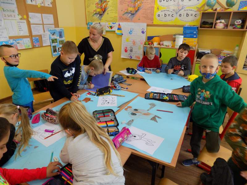 Na zdjęciu widać dzieci siedzące przy stolikach w trakcie kolorowania obrazków. W tle widać wychowawczynię klasy