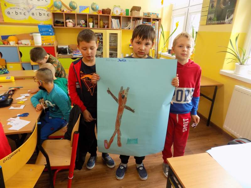 Na zdjęciu widać grupa trzech chłopców pokazuje wykonaną przez siebie pracę plastyczną na dużej karcie papieru. W tle widać innych uczniów siedzących przy stoliku