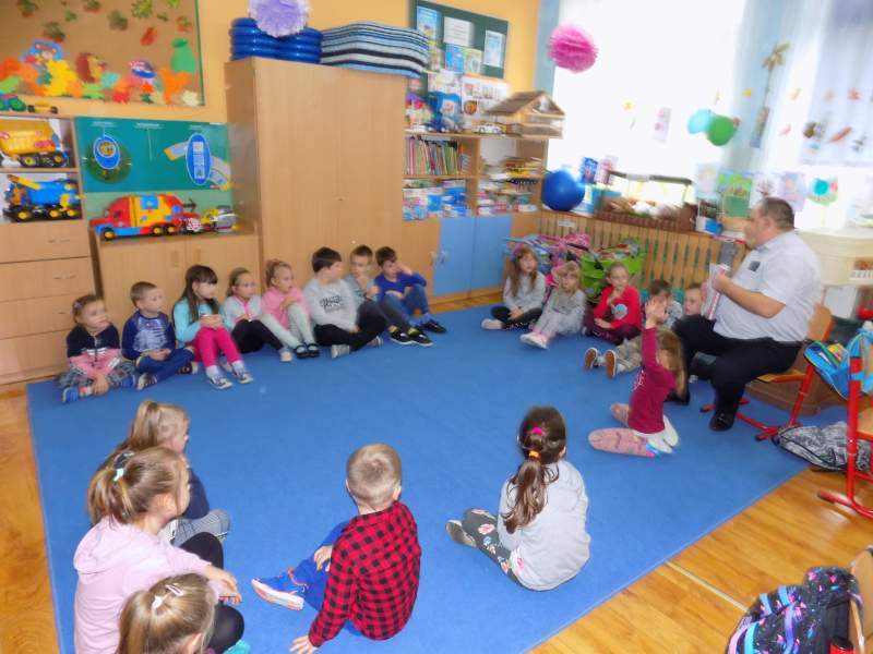 Na zdjęciu widać dzieci siedzące w kręgu w klasie na dywanie. W dali widać dyrektora biblioteki siedzącego na krześle i czytającego dzieciom bajki