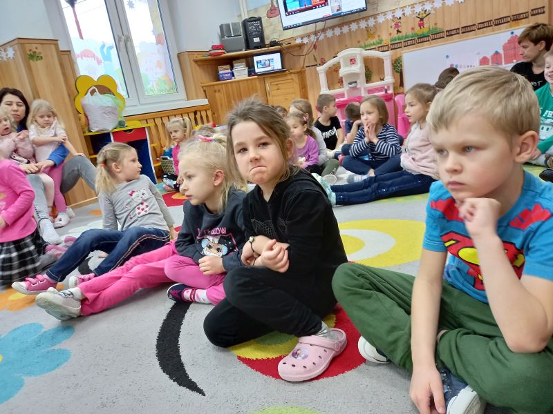 Zdjęcie przedstawia przedszkolaków siedzących na dywanie w przedszkolu