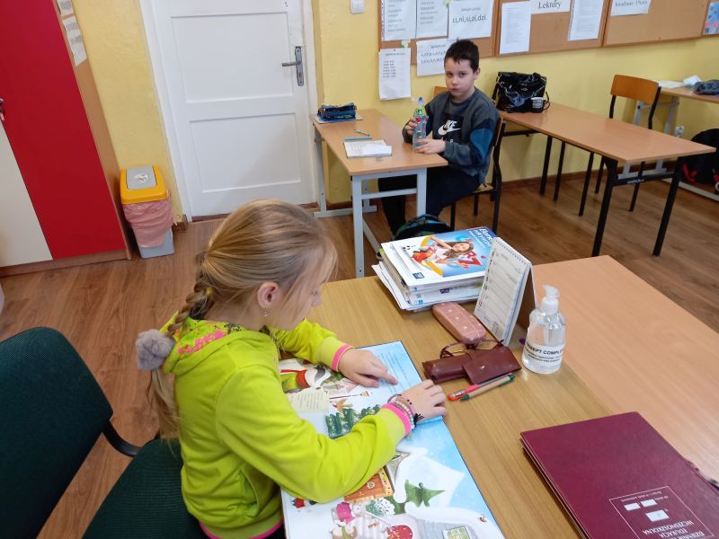 Zdjęcie przedstawia dziewczynkę siedzącą przy biurku nauczyciela w klasie i czytającą innym dzieciom siedzącym w ławkach bajkę o Mikołaju
