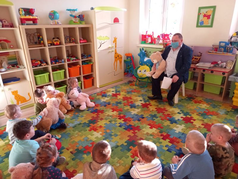 Zdjęcie przedstawia Dyrektora Biblioteki w trakcie zajęć z przedszkolakami. Dzieci siedzą na dywanie w kręgu wokół dyrektora. Dyrektor siedzi na krześle, w ręku trzyma pluszowego misia