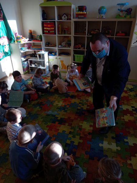 Zdjęcie przedstawia Dyrektora Biblioteki w trakcie zajęć z przedszkolakami. Dzieci siedzą na dywanie. Dyrektor biblioteki wręcza im kolorowe książeczki
