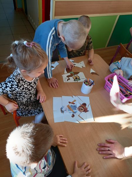 Zdjęcie przedstawia dzieci siedzące przy stoliku i układające puzzle z obrazkami misia