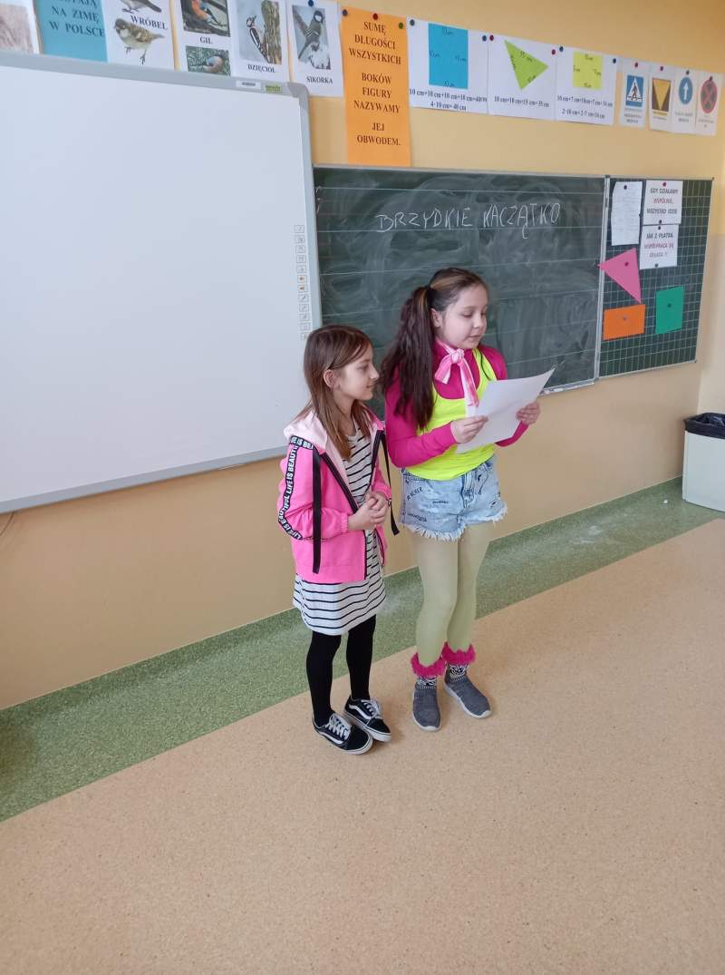Fotografia przedstawia dwie uczennice stojące w klasie przy tablicy. Uczennice czytają napisaną przez siebie bajkę